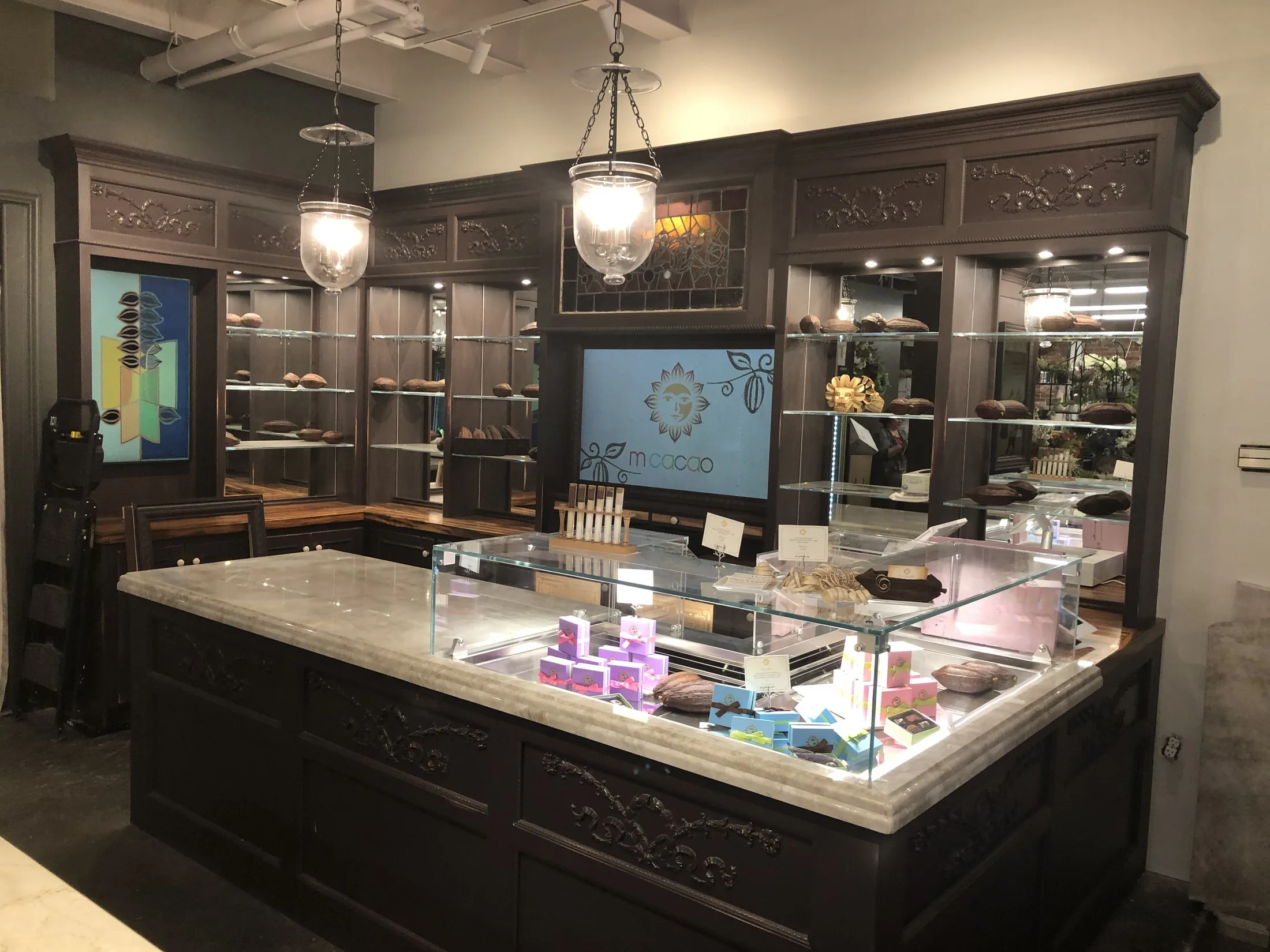 The m cacao retail room: a marble-topped display case in front of dark millwork, glass shelves, cacao pods, lighting, and the m cacao sign.